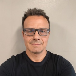 Head-and-shoulders portrait of a man with dark hair and blue glasses, wearing a black shirt against a gray background.
